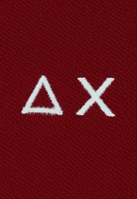 Lo sfondo tessile rosso presenta due simboli ricamati bianchi: un triangolo e una "X", con un aspetto morbido e texture.