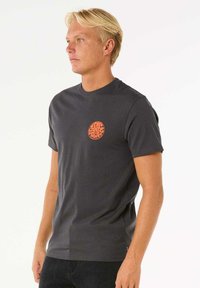 Graues Baumwoll-T-Shirt mit einem kleinen orangefarbenen runden Logo auf der linken Brustseite. Kurze Ärmel, Rundhalsausschnitt und glatte Textur.