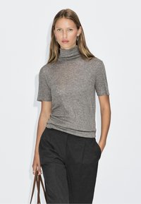 Light gray turtleneck top in a soft knit material, short sleeves, fitted design, worn with dark trousers, held with a brown shoulder strap.