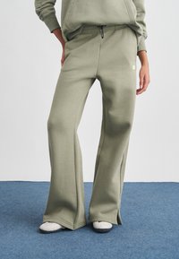 Άτομο φορώντας ελαφρώς χακί φαρδιά παντελόνια φόρμας και αντιστοιχισμένο φούτερ, στέκεται σε μπλε χαλί με το ένα χέρι στην τσέπη και λευκά παπούτσια ορατά.