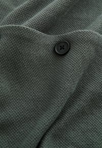 Tissu texturé vert foncé avec un seul bouton noir étiqueté "DOWNN TIME" cousu près du centre.