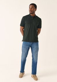 Ciemnozielona koszulka polo z krótkim rękawem i subtelnym logo na piersi, zestawiona z niebieskimi dżinsami i beżowym obuwiem. Neutralne tło.