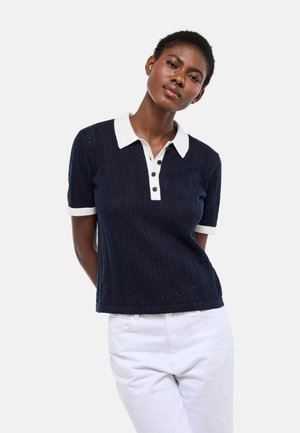Femme portant un polo en maille bleu marine à manches courtes avec col et poignets blancs, associé à un pantalon blanc, debout devant un fond uni.