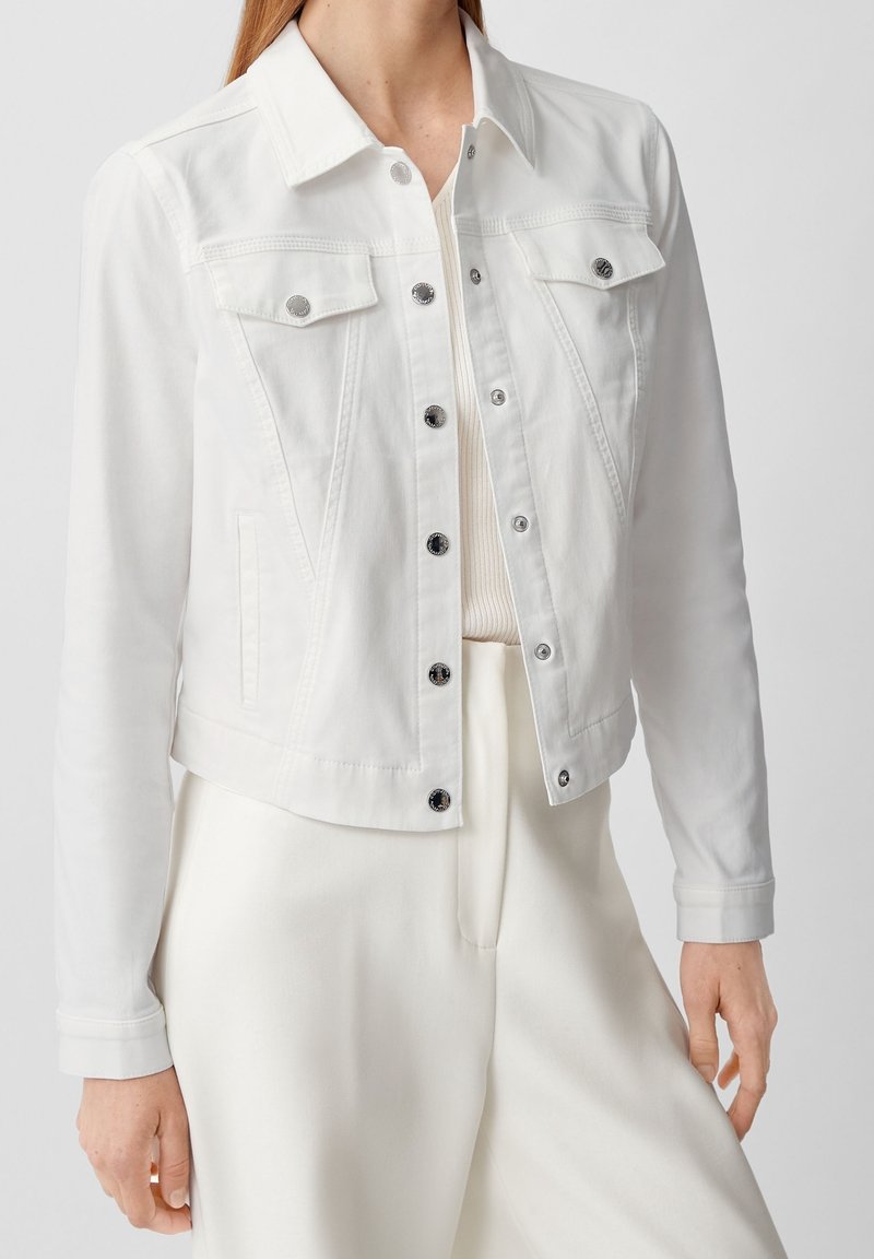 Femme portant une veste en jean blanche avec des boutons-pression métalliques sur un haut côtelé clair et un pantalon large crème, sur un fond uni.