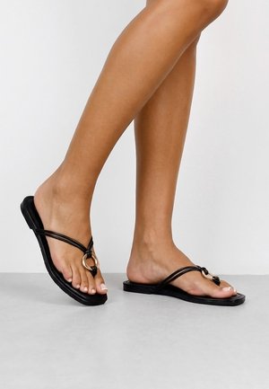 Piernas con sandalias negras sin cierre con tiras finas y un detalle decorativo de anillo dorado sobre un suelo gris claro y fondo blanco.