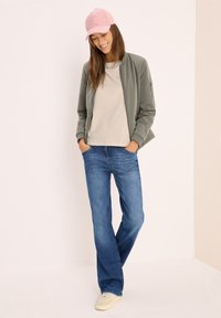 Lightweight olive green bomber jacket over a gray shirt, paired with blue flared jeans and cream sneakers. Pink corduroy cap adds accent.