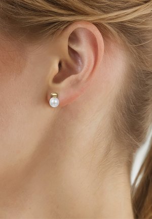 Primer plano de la oreja de una mujer que lleva un pendiente de oro con un perno coronado por una perla blanca redonda. Cabello rubio recogido detrás de la oreja.