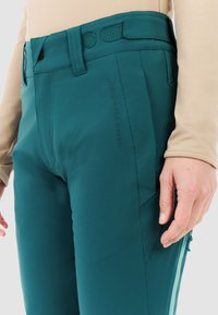 Tegengroen broek met een gladde, rekbare stof, een getailleerde pasvorm en een platte voorkant. Opvallende details zijn de riemlussen en een ritszak.