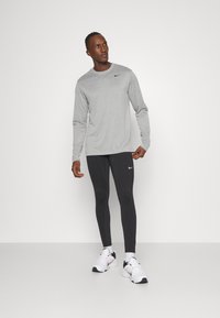 Chemise de sport à manches longues grise avec un logo noir, associée à un legging noir et des chaussures de sport blanches. Tissu lisse, design ajusté.