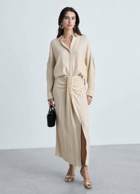 Beige shirt dress with a knotted waist detail and slit, made of soft fabric. Worn with gold sandals and holding a small black handbag.