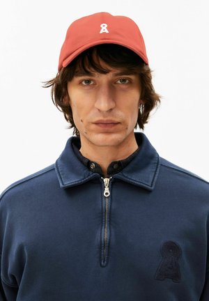 Junger Mann mit mittellangen dunklen Haaren, der eine rote Mütze mit weißem Logo und einen marineblauen Kapuzenpullover mit Reißverschluss und passendem Logopatch trägt.