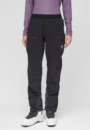 Personne portant un pantalon d'extérieur noir zippé, un t-shirt à manches longues violet et des chaussures de ski blanches, debout devant un fond uni.