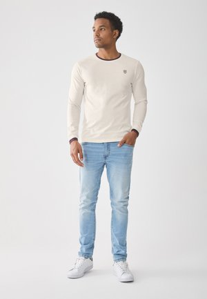 Langärmliges cremefarbenes Hemd mit einem kontrastierenden runden Ausschnitt, kombiniert mit hellblauen Jeans und weißen Turnschuhen. Dezentes Logo auf der Brust.
