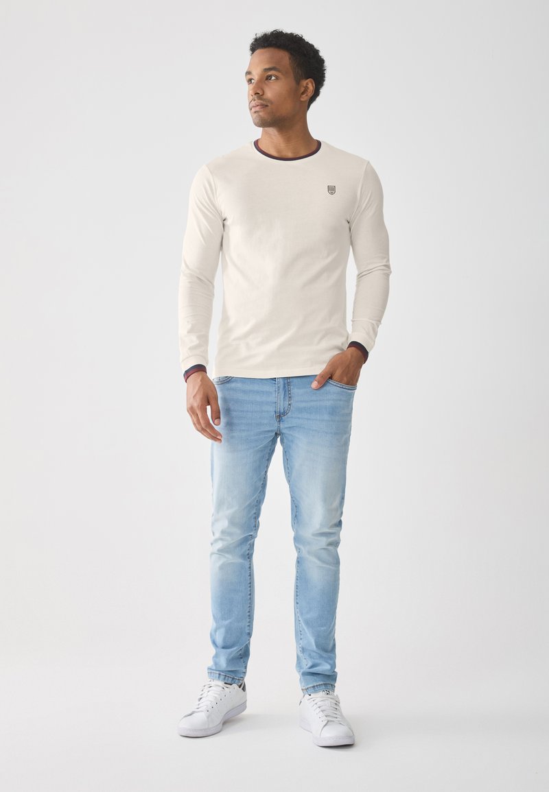 Camisa de manga larga color crema con un escote redondeado en contraste, combinada con jeans de mezclilla azul claro y zapatillas blancas. Sutil logo en el pecho.