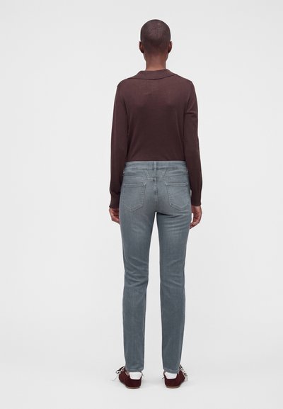 Personne aux cheveux courts portant une chemise marron à manches longues, un jean bleu clair, des chaussettes blanches et des chaussures rouge foncé, debout de dos.
