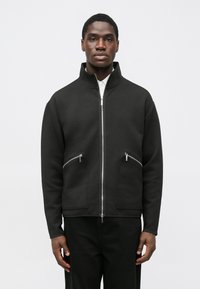 Black zip-up jacket in soft fabric, featuring a high collar, two front zippered pockets, and a relaxed fit. Smooth texture with minimal design.