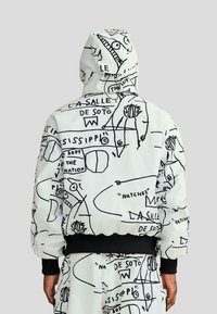 Fehér kapucnis dzseki fekete absztrakt mintákkal, rajzfilmszerű elemekkel és szöveggel. Fekete bordázott mandzsetták és szegély. Puha, párnázott textúra.