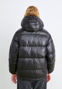 Čierna puffer bunda s kapucňou, s prešívaným dizajnom a lesklou, hladkou tkaninou. Stredne široký silueta s elastickými manžetami na zápästiach.