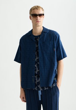 Giovane uomo che indossa occhiali da sole scuri e una camicia blu scuro a maniche corte sopra una camicia fantasia, con pantaloni blu scuro coordinati.