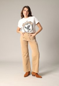 T-shirt graphique blanc avec un design d'aigle superposé sur un pantalon en velours côtelé beige, associé à des bottines en daim marron.