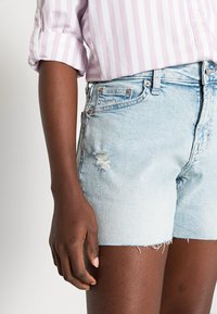 Ljusblå denimshorts med fransad nederkant, med slitna detaljer framtill och metallknäppning. Randig skjorta i tyg är delvis synlig.