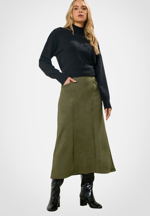 Blonde vrouw met een zwart trui met lange mouwen, een olijfgroene midirok met zakken en glanzende zwarte laarzen met hakken, staand met één hand in de zak.