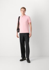 Muž ve růžovém polotriku, černých kalhotách, černých botách a černé crossbody tašce stojí před čistým bílým pozadím.