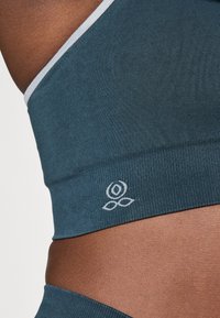Close-up de torso a usar um top desportivo sem emendas em tom de teal, com um pequeno logotipo floral branco perto da caixa torácica e acabamento branco na alça.