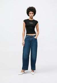 Zwart T-shirt met korte mouwen en wit logo, gecombineerd met losse blauwe spijkerbroek en witte schoenen met puntige neus. Eenvoudig ontwerp, casual pasvorm.