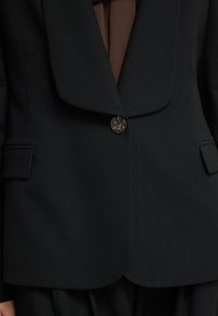 Zwarte pinstripe blazer met een brede revers, enkele knoopsluiting en twee frontzakken. Gemaakt van een gladde stof met een subtiele glans.