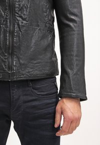 Particolare di una persona che indossa una giacca di pelle nera e jeans scuri, mostrando i dettagli della texture della giacca e la mano appoggiata di lato.