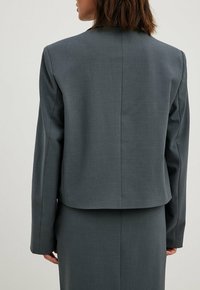 Blazer grigio aderente con spalline, maniche lunghe, cucitura posteriore e orlo corto. Realizzato in tessuto liscio e texture.