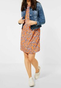 Denimjacka över en orange paisley-mönstrad klänning. Klänningen är knälång, har korta ärmar och en avslappnad passform. Vita sneakers.