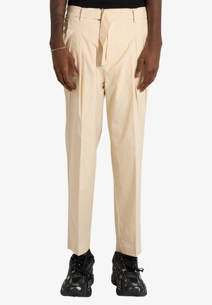 Uomo con pantaloni beige plissettati con cintura in tessuto e sneakers nere chunky, in piedi davanti a uno sfondo semplice.