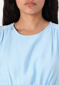 Ανοιχτό μπλε κοντομάνικο μπλουζάκι με στρογγυλή λαιμόκοψη, που διαθέτει συγκεντρωμένες λεπτομέρειες στους ώμους και ένα μαλακό, υφασμάτινο υλικό.