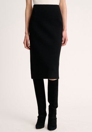 Femme portant une jupe crayon noire longueur genou associée à des bottes noires hautes jusqu'au genou et un haut blanc, debout devant un fond uni.