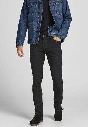 Homme portant une veste en jean bleu foncé, une chemise noire, un jean noir slim et des chaussures en daim noires, debout avec une main dans la poche.