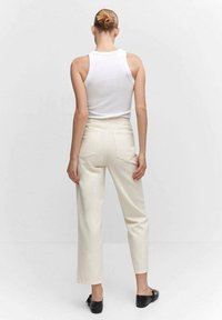 Mujer con el pelo recogido en un moño, vistiendo una blusa blanca sin mangas, pantalones de tiro alto en color blanco roto y zapatos planos negros, de pie frente a un fondo liso.