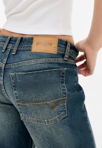 Blauwe spijkerbroek van denim met een leren label met "BDG JEANS" op de taille, oranje stiksels en een slim fit ontwerp met zichtbare achterzakken.