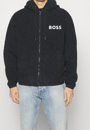 Mand iført sort fleece-zip-up-hættetrøje med "BOSS"-logo på brystet og lyseblå jeans, stående mod en ensfarvet baggrund.