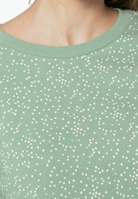 Haut en maille verte avec un motif texturé de petits carrés blancs, doté d'un col côtelé et d'un tissu doux. Vue en gros plan de la partie supérieure.