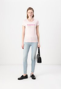 T-shirt ajusté rose clair avec le logo "GUESS" en dégradé de couleurs. Associé à un jean skinny bleu clair et des mocassins noirs, tenant un sac à main noir.