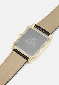 Montre carrée dorée avec un dos en acier inoxydable, bracelet en simili cuir noir et accents en cuir beige lisse. Marquée avec les détails de la marque et du modèle.
