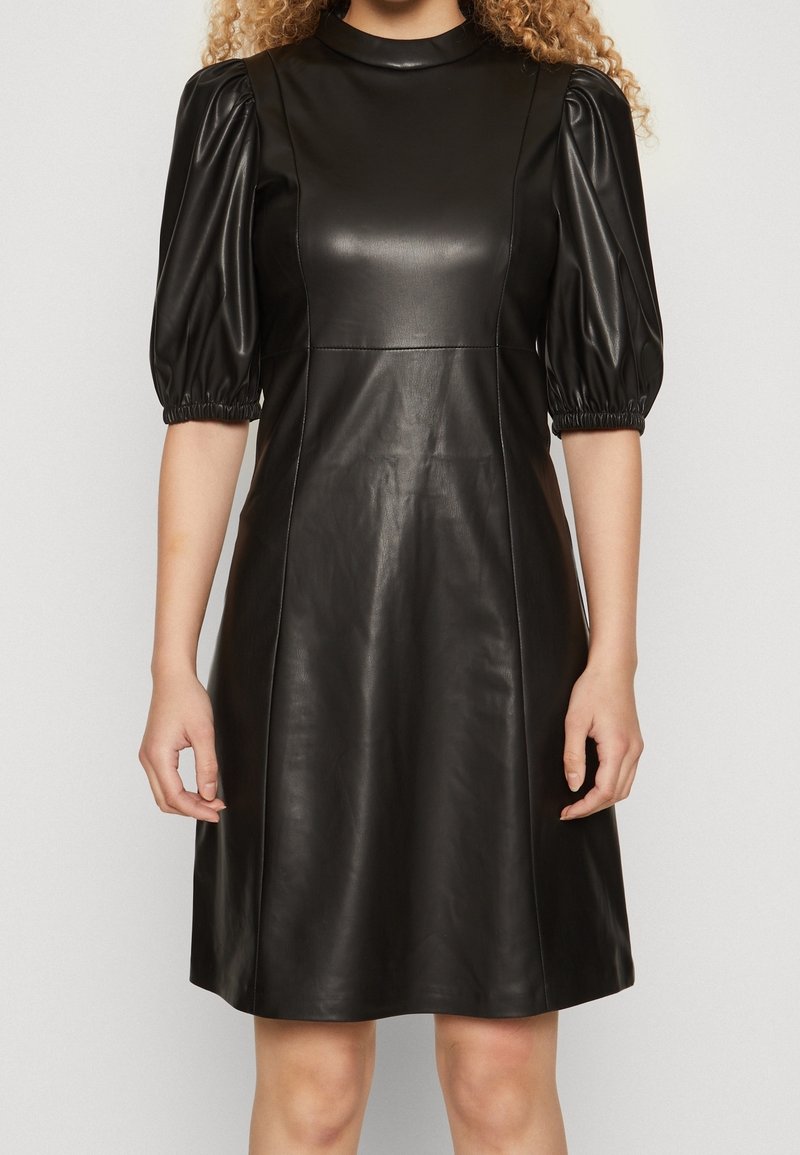 Robe noire en similicuir longueur genou avec manches bouffantes aux coudes et col rond haut, portée par une femme debout devant un fond uni.