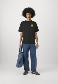 Camiseta negra de manga corta con un gráfico colorido en el pecho, combinada con unos jeans de mezclilla azules sueltos y zapatillas marrones. Sosteniendo una bolsa de mano azul.