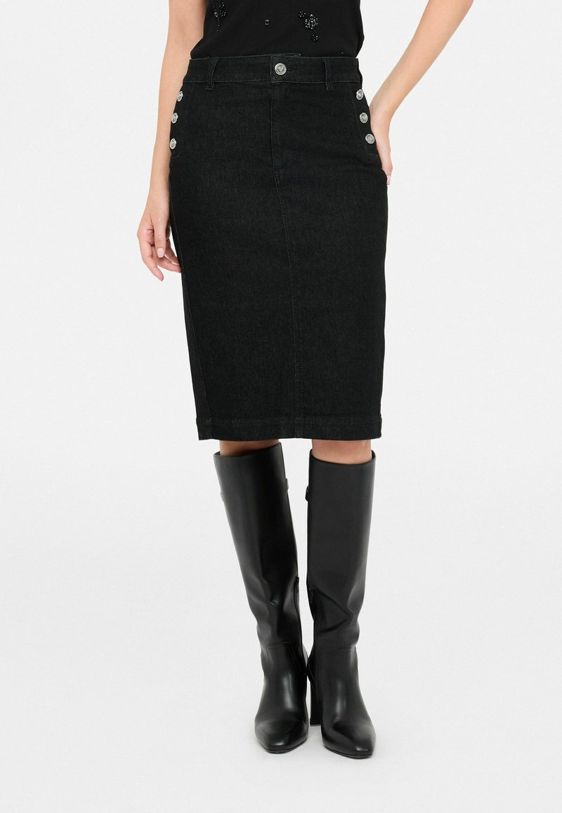 Femme portant une jupe crayon noire longueur genou avec des boutons latéraux, associée à des bottes en cuir noires montantes, debout sur un fond blanc.
