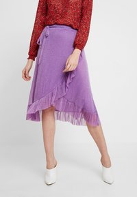 Fialová průsvitná sukně s volánkovým lemem a zavazováním v pase. Nošena s červeným vzorovaným topem. Obuv jsou bílé otevřené podpatky. Jednoduché pozadí.