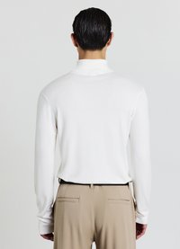 Maglione bianco a collo alto in maglia con maniche lunghe, texture liscia e colletto a coste, abbinato a pantaloni beige su misura con tasche posteriori.