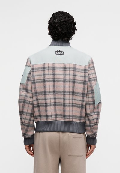 Homme aux cheveux bouclés portant une veste à carreaux rose et grise avec des patches bleu clair sur les épaules et des poignets et une ceinture côtelés gris.