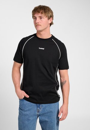 Mężczyzna z wąsami w czarnej koszulce hummel z białymi lamówkami i niebieskich dżinsach, stojący z jedną ręką w kieszeni na białym tle.